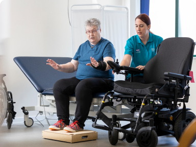 A photograph of a service user and one of our clinicians in clinic. The service user is sat on a plinth and is discussing her power wheelchair with the wheelchair service clinician.